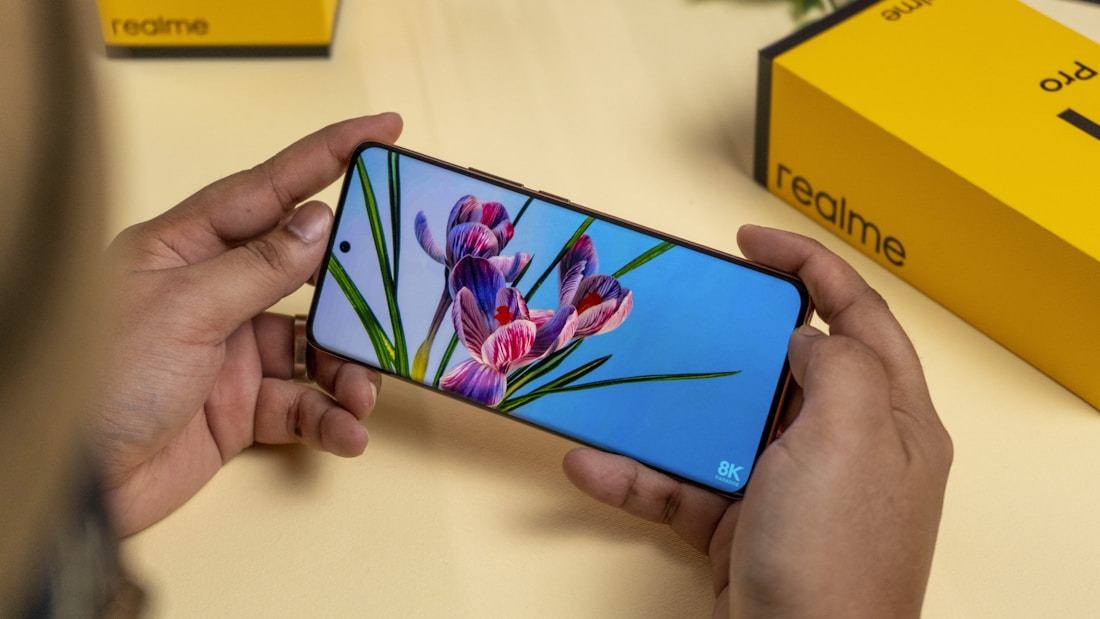 A person holds a smartphone displaying an image of flowers with a vivid screen resolution. A yellow and black Realme product box is visible in the background on the table.
