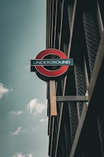 A vintage-style sign with the word 'Underground' in white letters on a blue background, encircled by a red and black roundel, mounted on the side of a building with a metal and glass facade. The sky is overcast with some clouds.