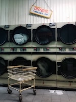 A row of industrial dryers with a sign above them indicating the cost and time for use. Several of the dryers are running, visible by the spinning clothes inside. A metal laundry cart is positioned in front of the dryers, and the floor is tiled.