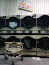 A row of industrial dryers with a sign above them indicating the cost and time for use. Several of the dryers are running, visible by the spinning clothes inside. A metal laundry cart is positioned in front of the dryers, and the floor is tiled.