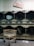A row of industrial dryers with a sign above them indicating the cost and time for use. Several of the dryers are running, visible by the spinning clothes inside. A metal laundry cart is positioned in front of the dryers, and the floor is tiled.