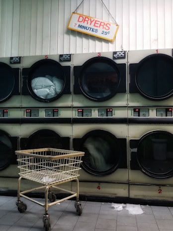 A row of industrial dryers with a sign above them indicating the cost and time for use. Several of the dryers are running, visible by the spinning clothes inside. A metal laundry cart is positioned in front of the dryers, and the floor is tiled.