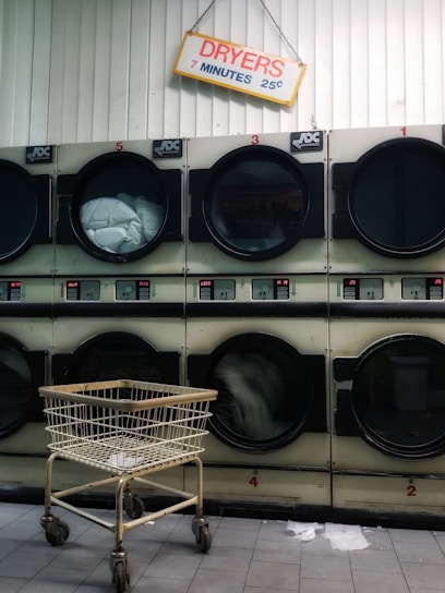 A row of industrial dryers with a sign above them indicating the cost and time for use. Several of the dryers are running, visible by the spinning clothes inside. A metal laundry cart is positioned in front of the dryers, and the floor is tiled.