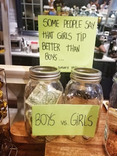 Two large glass jars labeled 'BOYS' and 'GIRLS' are placed on a wooden counter, with a note above them suggesting that girls tip better than boys. The jars appear to be tip jars for a survey, with money visible in at least one of them. The background suggests a café setting with people and equipment visible.