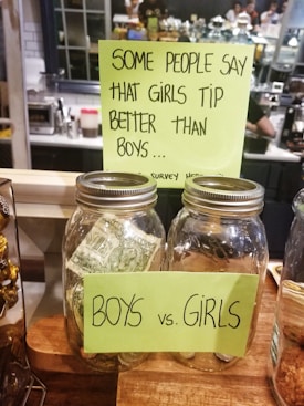 Two large glass jars labeled 'BOYS' and 'GIRLS' are placed on a wooden counter, with a note above them suggesting that girls tip better than boys. The jars appear to be tip jars for a survey, with money visible in at least one of them. The background suggests a caf&eacute; setting with people and equipment visible.