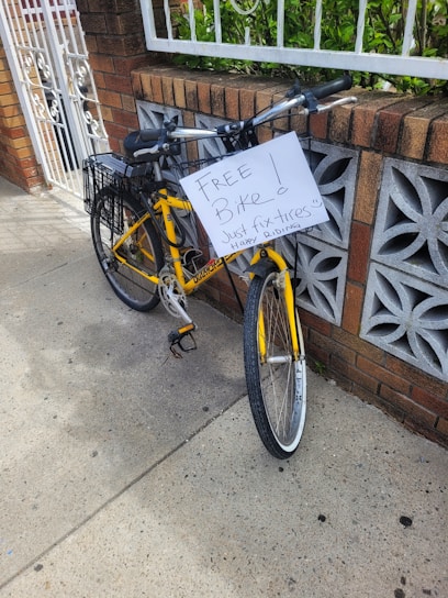 A yellow bicycle with a basket is parked on a sidewalk, leaning against a decorative brick wall. A handwritten sign on the bicycle reads 'FREE Bike! Just fix tires. Happy riding.' The scene includes a white metal gate and some greenery in the background.
