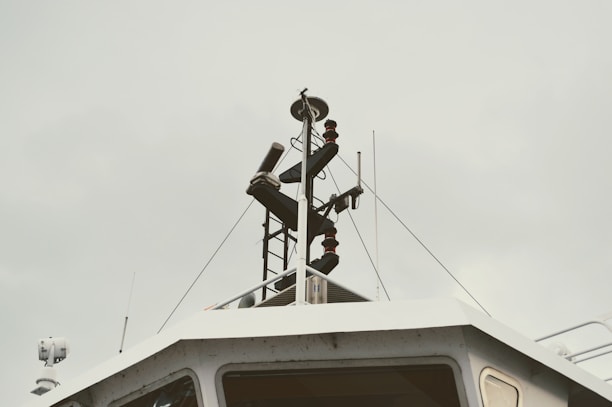 A detailed view of the upper part of a boat or ship's structure, featuring antennas, radar equipment, and other navigational instruments. The background sky appears cloudy and overcast.