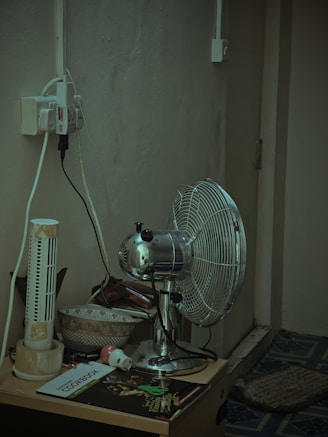 A corner of a room featuring a metallic oscillating fan placed on a small table. Beside the fan, there is an old, damaged tower fan. The table also holds a couple of bowls, a cookbook, a small electronic plug, and various other items. There are multiple electrical cords plugged into an outlet that is mounted on the wall. The wall and the carpeted floor appear worn and aged.