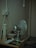 A corner of a room featuring a metallic oscillating fan placed on a small table. Beside the fan, there is an old, damaged tower fan. The table also holds a couple of bowls, a cookbook, a small electronic plug, and various other items. There are multiple electrical cords plugged into an outlet that is mounted on the wall. The wall and the carpeted floor appear worn and aged.