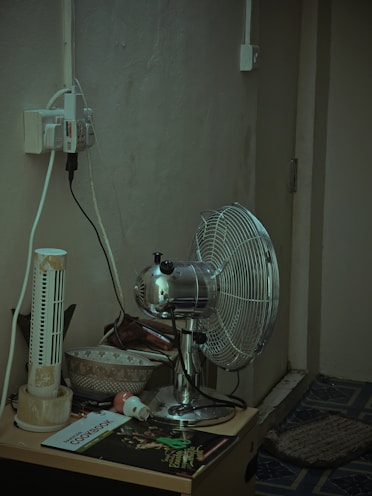 A corner of a room featuring a metallic oscillating fan placed on a small table. Beside the fan, there is an old, damaged tower fan. The table also holds a couple of bowls, a cookbook, a small electronic plug, and various other items. There are multiple electrical cords plugged into an outlet that is mounted on the wall. The wall and the carpeted floor appear worn and aged.
