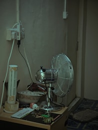 A corner of a room featuring a metallic oscillating fan placed on a small table. Beside the fan, there is an old, damaged tower fan. The table also holds a couple of bowls, a cookbook, a small electronic plug, and various other items. There are multiple electrical cords plugged into an outlet that is mounted on the wall. The wall and the carpeted floor appear worn and aged.