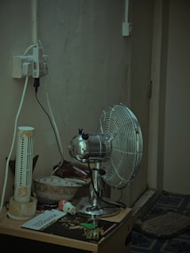 A corner of a room featuring a metallic oscillating fan placed on a small table. Beside the fan, there is an old, damaged tower fan. The table also holds a couple of bowls, a cookbook, a small electronic plug, and various other items. There are multiple electrical cords plugged into an outlet that is mounted on the wall. The wall and the carpeted floor appear worn and aged.
