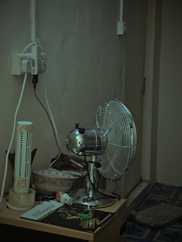 A corner of a room featuring a metallic oscillating fan placed on a small table. Beside the fan, there is an old, damaged tower fan. The table also holds a couple of bowls, a cookbook, a small electronic plug, and various other items. There are multiple electrical cords plugged into an outlet that is mounted on the wall. The wall and the carpeted floor appear worn and aged.