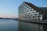 A modern, stepped apartment building with numerous balconies reflects the evening light beside a calm body of water. The architecture is unique, resembling a staircase with glass railings. There are a few people walking along the waterside promenade.