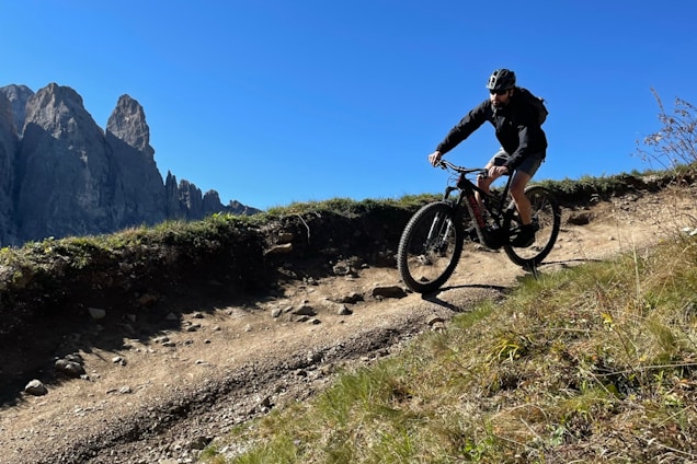 A rider powering down a rugged, rocky trail with dense forest surrounding the path under a clear blue sky.