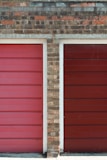 Two adjacent garage doors with horizontal panels, framed by a brick wall. The colors of the doors are distinct shades of red, with the left one being brighter. The brick wall has a mix of shades, including brown and gray, with visible mortar lines.