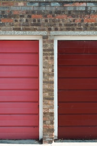 Two adjacent garage doors with horizontal panels, framed by a brick wall. The colors of the doors are distinct shades of red, with the left one being brighter. The brick wall has a mix of shades, including brown and gray, with visible mortar lines.