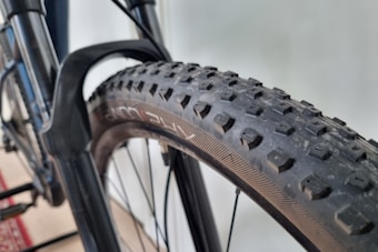 Close-up view of a bicycle tire with a textured tread pattern, mounted on a metal frame. The tire appears robust and is likely designed for off-road use. The background is slightly blurred, suggesting a focus on the bicycle component.