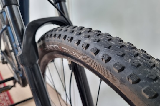 Close-up view of a bicycle tire with a textured tread pattern, mounted on a metal frame. The tire appears robust and is likely designed for off-road use. The background is slightly blurred, suggesting a focus on the bicycle component.