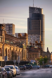 A cityscape featuring a large, modern skyscraper in the background with the word 'UNIQA' displayed at the top. In the foreground, a row of historic, ornate brick buildings with arched windows lines the street. Parked cars are visible along the street, and there are trees and street signs along the sidewalk. The lighting suggests a late afternoon or early evening setting with golden hues illuminating the scene.