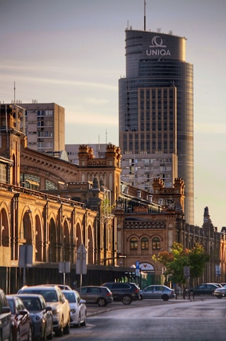 A cityscape featuring a large, modern skyscraper in the background with the word 'UNIQA' displayed at the top. In the foreground, a row of historic, ornate brick buildings with arched windows lines the street. Parked cars are visible along the street, and there are trees and street signs along the sidewalk. The lighting suggests a late afternoon or early evening setting with golden hues illuminating the scene.