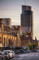A cityscape featuring a large, modern skyscraper in the background with the word 'UNIQA' displayed at the top. In the foreground, a row of historic, ornate brick buildings with arched windows lines the street. Parked cars are visible along the street, and there are trees and street signs along the sidewalk. The lighting suggests a late afternoon or early evening setting with golden hues illuminating the scene.
