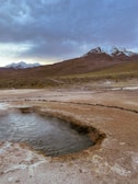 A geothermal landscape with a steaming pool surrounded by rocky terrain under a cloudy sky.