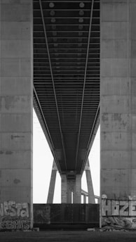 An architectural view of a large concrete bridge captured from underneath, showcasing its massive support columns and intricate underside structure. Graffiti adorns the lower parts of the columns, adding an urban art element to the industrial setting.