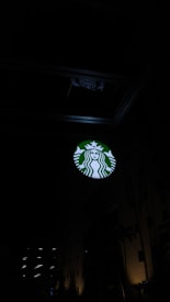 A lit circular sign featuring a mermaid logo is prominently displayed against a dark background. The sign is green and white, and it is positioned on a tall building. Some architectural details of the structure are visible, with faint lights coming from some windows.