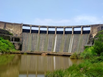 A large concrete dam sits in the background with water cascading down its surface. The sky is a clear blue, and green vegetation surrounds the dam, especially on the right side. In the foreground, there is a calm body of water reflecting parts of the dam and the nearby greenery.