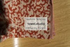 A close-up of a towel with a textured red and beige pattern is placed on a wooden surface. A label is sewn onto the towel, displaying the text 'Designed in Berlin Made in Germany towel.studio 100% Organic Cotton' along with care symbols.