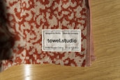 A close-up of a towel with a textured red and beige pattern is placed on a wooden surface. A label is sewn onto the towel, displaying the text 'Designed in Berlin Made in Germany towel.studio 100% Organic Cotton' along with care symbols.