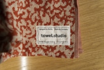 A close-up of a towel with a textured red and beige pattern is placed on a wooden surface. A label is sewn onto the towel, displaying the text 'Designed in Berlin Made in Germany towel.studio 100% Organic Cotton' along with care symbols.