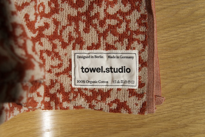 A close-up of a towel with a textured red and beige pattern is placed on a wooden surface. A label is sewn onto the towel, displaying the text 'Designed in Berlin Made in Germany towel.studio 100% Organic Cotton' along with care symbols.