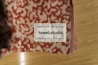 A close-up of a towel with a textured red and beige pattern is placed on a wooden surface. A label is sewn onto the towel, displaying the text 'Designed in Berlin Made in Germany towel.studio 100% Organic Cotton' along with care symbols.