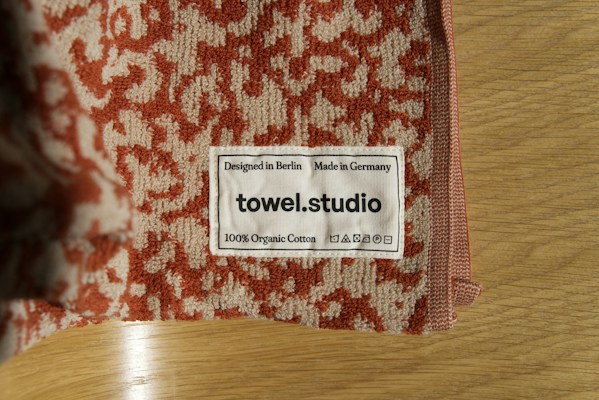 A close-up of a towel with a textured red and beige pattern is placed on a wooden surface. A label is sewn onto the towel, displaying the text 'Designed in Berlin Made in Germany towel.studio 100% Organic Cotton' along with care symbols.