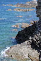 The image features a rugged coastline with large, dark rocks jutting out into a vibrant blue ocean. Gentle waves lap against the rocks, creating a contrast between the rough textures of the stone and the smooth expanse of the water. The scene conveys a sense of natural beauty and tranquility.
