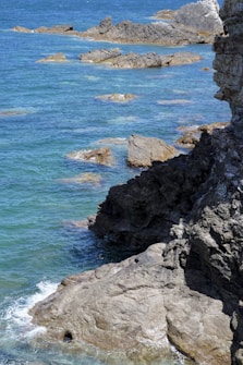 The image features a rugged coastline with large, dark rocks jutting out into a vibrant blue ocean. Gentle waves lap against the rocks, creating a contrast between the rough textures of the stone and the smooth expanse of the water. The scene conveys a sense of natural beauty and tranquility.