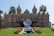 A grand historic building with ornate architecture, two palm trees framing the front, a reflective sculpture in the center, and a manicured lawn with red flowers. The façade features detailed carvings, columns, and statues.