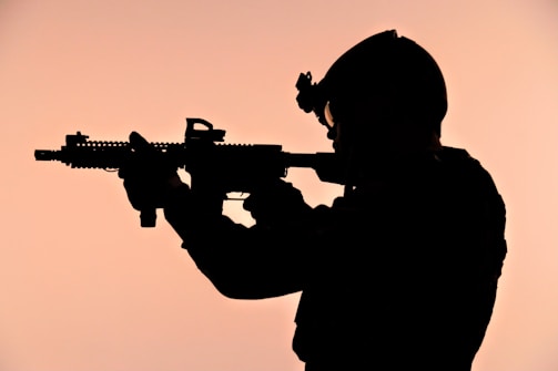 A silhouette of a person in military gear aiming a rifle against a softly glowing background. The figure is wearing a helmet and tactical equipment, standing out starkly due to the contrast.