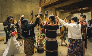 A group of people dressed in traditional and colorful clothing are participating in what appears to be a cultural dance or ceremony indoors. The room has a rustic decor with wooden beams and earthen walls. The individuals are holding hands in a circle around some traditional artifacts and musical instruments placed in the center.