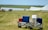 Two carts filled with various luggage, including red, blue, and white suitcases, are positioned on a grassy field. In the background, a body of water and some tents can be seen, suggesting a camping or outdoor event.