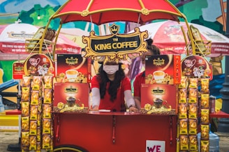 A vibrant outdoor setup featuring a King Coffee kiosk with an eye-catching red and yellow theme. A woman wearing a red t-shirt and a face mask is standing at the kiosk surrounded by various coffee product displays. The kiosk is covered with decorative banners and an umbrella, and more products and promotional materials are visible, creating a lively and colorful market atmosphere.