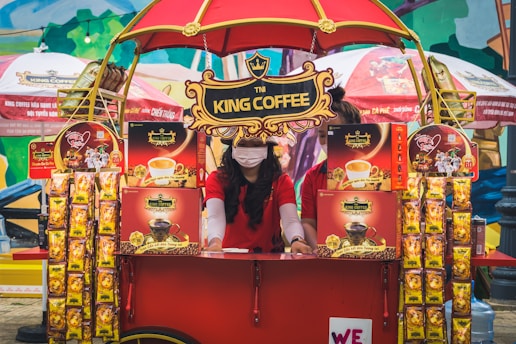 A vibrant outdoor setup featuring a King Coffee kiosk with an eye-catching red and yellow theme. A woman wearing a red t-shirt and a face mask is standing at the kiosk surrounded by various coffee product displays. The kiosk is covered with decorative banners and an umbrella, and more products and promotional materials are visible, creating a lively and colorful market atmosphere.