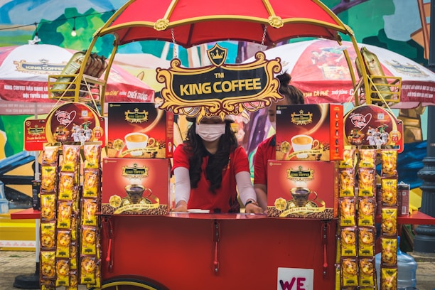 A vibrant outdoor setup featuring a King Coffee kiosk with an eye-catching red and yellow theme. A woman wearing a red t-shirt and a face mask is standing at the kiosk surrounded by various coffee product displays. The kiosk is covered with decorative banners and an umbrella, and more products and promotional materials are visible, creating a lively and colorful market atmosphere.