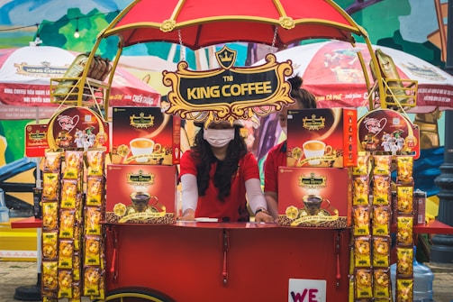A vibrant outdoor setup featuring a King Coffee kiosk with an eye-catching red and yellow theme. A woman wearing a red t-shirt and a face mask is standing at the kiosk surrounded by various coffee product displays. The kiosk is covered with decorative banners and an umbrella, and more products and promotional materials are visible, creating a lively and colorful market atmosphere.