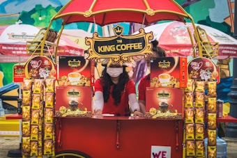 A vibrant outdoor setup featuring a King Coffee kiosk with an eye-catching red and yellow theme. A woman wearing a red t-shirt and a face mask is standing at the kiosk surrounded by various coffee product displays. The kiosk is covered with decorative banners and an umbrella, and more products and promotional materials are visible, creating a lively and colorful market atmosphere.