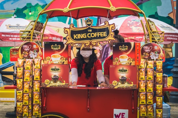 A vibrant outdoor setup featuring a King Coffee kiosk with an eye-catching red and yellow theme. A woman wearing a red t-shirt and a face mask is standing at the kiosk surrounded by various coffee product displays. The kiosk is covered with decorative banners and an umbrella, and more products and promotional materials are visible, creating a lively and colorful market atmosphere.