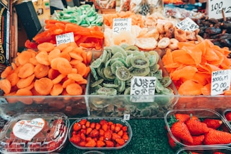 A vibrant market stall displaying an assortment of dried fruits, such as apricots, kiwis, and mangos. Fresh strawberries are also visible in plastic containers. Each section has price tags indicating the cost of the items.