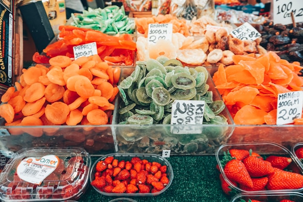 A vibrant market stall displaying an assortment of dried fruits, such as apricots, kiwis, and mangos. Fresh strawberries are also visible in plastic containers. Each section has price tags indicating the cost of the items.